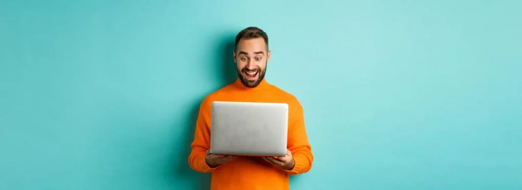 Uśmiechnięty mężczyzna w pomarańczowym swetrze korzysta z laptopa na tle turkusowej ściany – praca online, technologia, zadowolenie użytkownika.