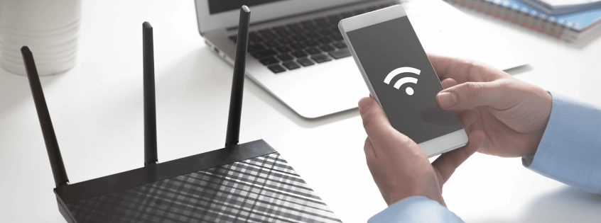 Osoba trzymająca smartfon z ikoną Wi-Fi obok routera bezprzewodowego i laptopa na biurku.